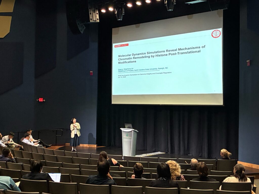 Rina Li on a stage in front of a pull down screen that has their presentation projected onto it. The slide is the title page that with teh title "Molecular dynamics simulations reveal mechanisms of chromatin remodeling by histone post-translational modifications." Li is in front of a crowd.